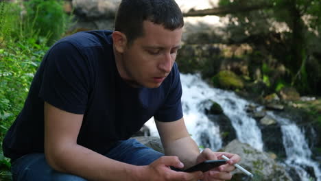 Young-alone-man-browsing-social-media-on-a-phone-and-smoking-cigarette-near-a-waterfall
