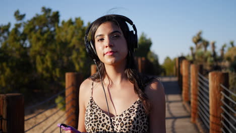 A-cute-young-hispanic-woman-listening-to-pop-music-on-her-headphones-and-smartphone-while-walking-in-slow-motion-outdoors-in-sunlight