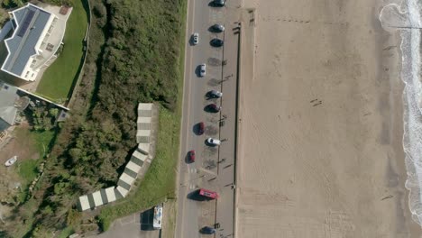Top-down-aerial-over-a-sandy-beach-connected-by-a-coast-road-and-a-car-park