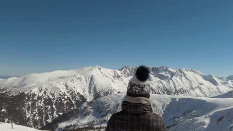 Girl-with-winter-hat-at-a-top-of-a-ski-slope,-looking-at-a-horizon-of-mountains-covered-with-snow