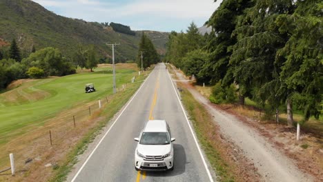 Luxury-suv-car-driving-along-golf-course-in-Arrowtown,-New-Zealand-while-golfers-crossing-road---Aerial