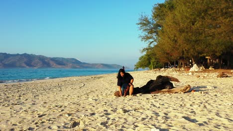 Girl-sitting-on-a-trunk-playing-with-white-sand-of-exotic-beach-washed-by-blue-sea-surrounding-tropical-island-in-Indonesia