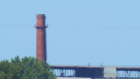 Distant-view-of-abandoned-metal-melting-factory-red-brick-chimney-in-sunny-summer-day-without-clouds,-medium-shot-from-a-distance