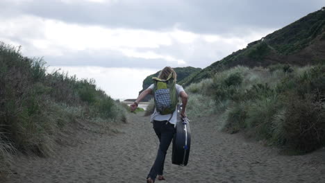 Slow-motion-of-a-young-blonde-man-running-happily-towards-the-ocean,-with-a-backpack-and-a-guitar-cover,-on-a-sandy-walkway-surrounded-by-high-grass-at-Bethells-Beach,-on-a-cloudy-and-windy-day