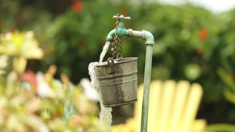 Water-from-a-garden-spout-pours-and-spills-over-a-hanging-bucket-into-a-larger-barrel-with-colored-lounge-chairs-in-the-background