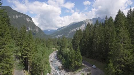 A-drone-flies-over-a-glacier-melt-river-bridge-and-valley-surrounded-by-a-high-mountains-and-a-lush-green-alpine-forest