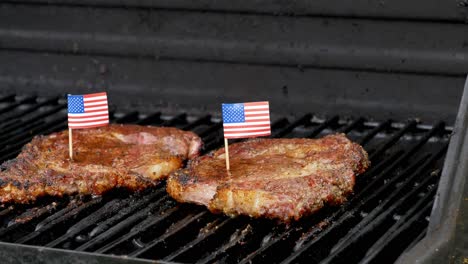 Eine-Wunderbare-Aufnahme-Von-Zwei-Saftigen-Rib-Eye-Steaks,-Die-Auf-Dem-Grill-Sitzen-Und-Mit-Zwei-Kleinen-Amerikanischen-Flaggen-Darauf-Mit-Zahnstochern-Grillen