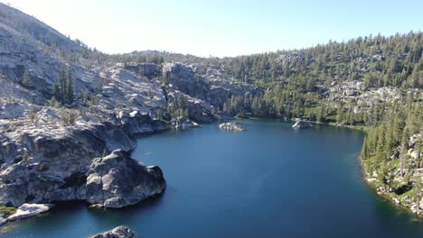 High-resolution-4K-drone-shot-circle-around-an-alpine-clear-water-lake-surrounded-by-trees,-rocks,-cliffs-in-the-California-wilderness-perfect-for-camping,-hiking,-and-backpacking-at-sunset-in-summer