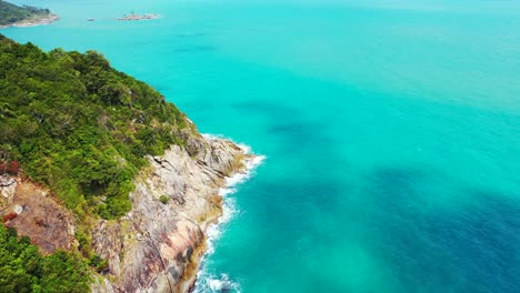 Beautiful-shoreline-of-tropical-island-with-lush-vegetation,-rocky-coast-splashed-by-waves-of-turquoise-sea-in-Thailand