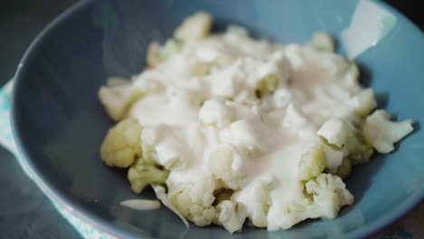Pouring-creamy-cheese-on-cooked-cauliflower-in-a-gray-plate-on-dark-background,-unconventional-healthy-alternative-to-Slovak-traditional-food-bryndzove-halusky