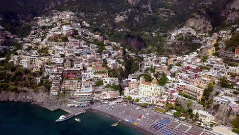 Positano-town-in-the-Amalfi-coast-of-Italy-with-boat-and-crowded-beach-umbrellas,-Aerial-pan-left-approach-shot