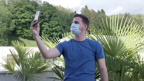 Young-man-in-blue-shirt-makes-selfies-in-the-garden-while-wearing-his-face-mask