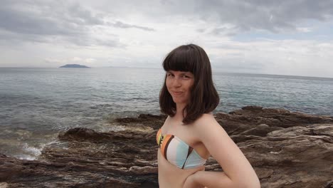 A-young-brunette-woman-with-bangs-smiling-and-having-fun-at-the-camera-on-a-rocky-beach-in-Greece-on-a-sunny-day