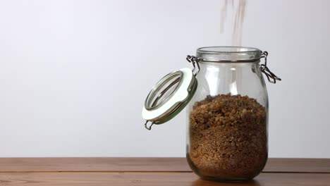 Cereal-being-poured-into-a-glass-jar-on-a-wooden-table-with-a-white-background
