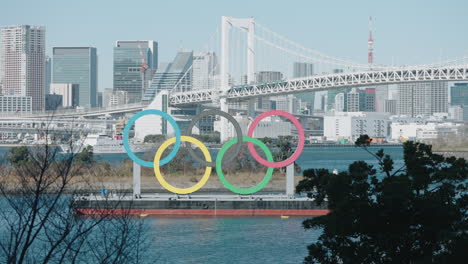 Olympic-Rings-Monument-At-Odaiba-Marine-Park-With-Sailing-Boat,-Cityscape,-Rainbow-Bridge-And-Tokyo-Tower-In-Background-In-Minato,-Tokyo,-Japan