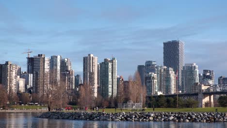 Wide-shot-of-a-small-park-by-the-sea-with-people-strolling-by-and-highrise-buildings-in-the-background-on-a-sunny-winter-day-in-Vancouver,-Canada
