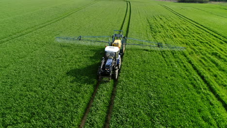 Drone-shot-of-a-blue-tractor-with-yellow-trailer-spraying-crops-on-green-field-with-insecticides