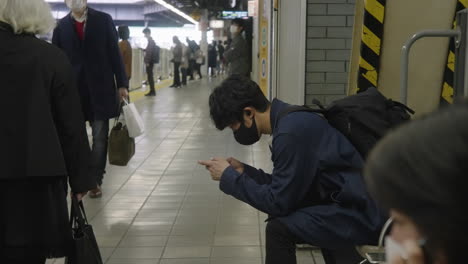 Hombre-Sentado-Usando-Un-Teléfono-Móvil-Con-Mascarilla-En-La-Plataforma-De-La-Estación-De-Tren-Durante-La-Pandemia-En-Tokio,-Japón
