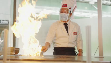 Teppanyaki-Show-In-Front-Of-Customers---Skilled-Chef-Wearing-Facemask-And-Face-Shield-With-Scraper-Puts-Oil-On-Fire-In-Hot-Griddle-In-An-Asian-Restaurant