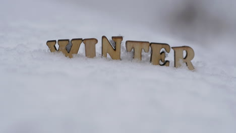 Das-Wort-Winter-Mit-Holzbuchstaben-Auf-Schnee-Geschrieben,-Der-Rack-Fokus-Ist-Nach-Oben-Geneigt
