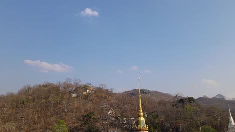 Wat-Phra-Phutthabat,-Saraburi,-Tailandia,-Toma-Aérea-Ascendente-Del-Templo-Budista-Dorado,-Deslizándose-Hacia-La-Izquierda-Revelando-árboles-Marrones-En-Verano,-Una-Colina-Con-Una-Escalera-Que-Sube-A-Otro-Templo