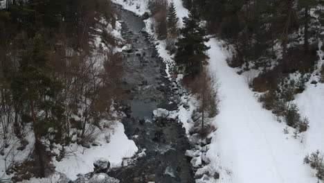 Aerial-view-of-small-river-in-rural-countryside-on-snowing-winter-day,-drone-shot