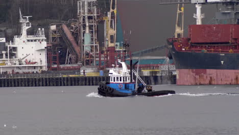Tracking-shot-of-tug-boat-cruising-on-river-westbound-with-gigantic-container-ship-with-freight-in-background