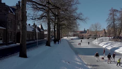 Aerial-snow-landscape-of-frozen-over-river-with-medieval-Berkelpoort-boat-portal-in-the-background-and-people-ice-skating-and-enjoying-the-solid-canal-against-a-blue-sky