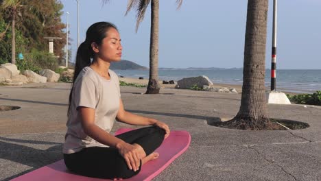 Calm-Asian-Woman-In-Casual-Sportswear-Practicing-Yoga-By-The-Shore,-Sitting-In-Lotus-Position-On-A-Yoga-Mat