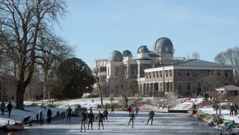 An-Einem-Sonnigen-Wintertag-Während-Der-Pandemie-Laufen-Menschen-Auf-Einem-Zugefrorenen-Kanal-Vor-Dem-Leiden-Observatorium-In-Den-Niederlanden-Schlittschuh
