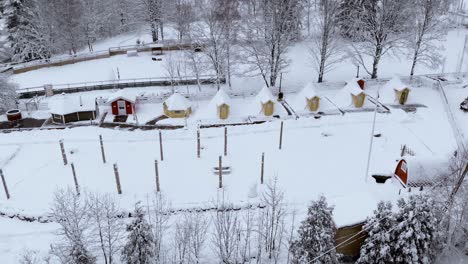 Aerial-view-over-cottages,-sauna-and-a-big-tent-in-the-Kalevala-Finnish-village-resort,-in-middle-of-snowy-forest,-cloudy,-winter-day---tilt-up,-drone-shot