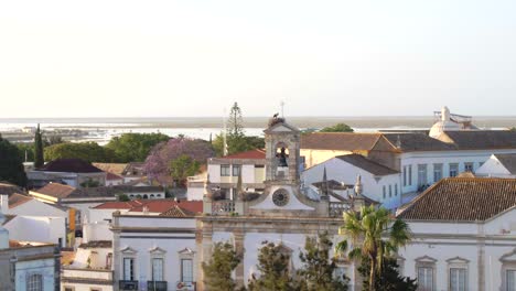 Dando-Vueltas-Alrededor-Del-Antiguo-Campanario-De-La-Iglesia-Expuesto-Rodeado-De-Edificios-Tradicionales-Europeos-Antiguos-En-Faro-Portugal,-Antena