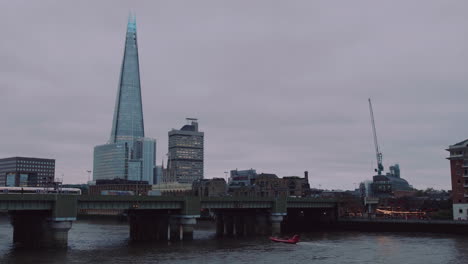 A-wide-shot-of-the-Shard-and-its-bankside-with-a-pub,-a-train-leaving-and-boats-passing