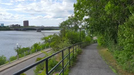 Walking-and-biking-trail-with-view-to-street-and-river-below-plus-bridge-and-buildings-in-background-under-blue-sky-in-summer-in-Ottawa-Ontario-Canada