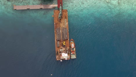 Crane-loading-pipes-on-flat-industrial-boat-surrounded-by-clear-transparent-indian-ocean-with-coral-reefs-in-Indonesia