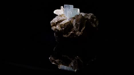An-aquamarine-crystal-emerging-from-the-matrix-of-muscovite-turns-slowly-with-a-reflection-of-the-sample-apparent-in-the-glass-base