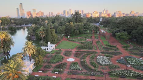 Aerial-jib-up-over-Rosedal-gardens-near-pond-in-Palermo-neighborhood,-skyline-in-background-at-sunset,-Buenos-Aires