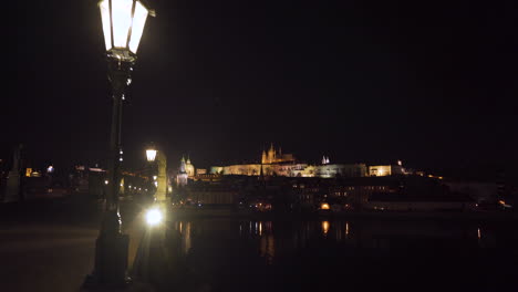 A-view-of-the-famous-Prague-castle-above-the-Vltava-river-from-Charles-bridge-at-night,-during-a-Covid-19-lockdown-with-no-people-anywhere,-lanterns-and-statues-on-the-bridge,-zoom-out-4k-shot
