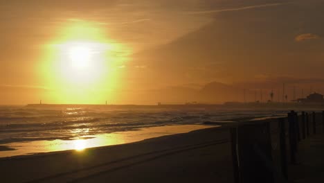 Hazy-sunrise-over-sandy-beach-and-harbor,-waves-rolling-in,-oliva,-mediterranean-coast,-spain