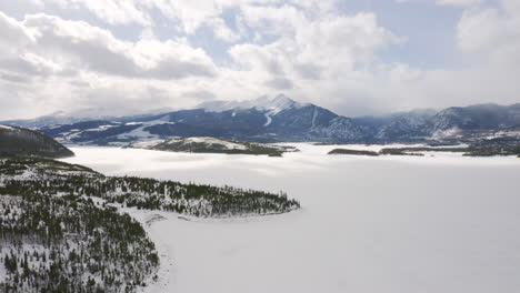 Die-Rückwärts-Bewegte-Luftaufnahme-Zeigt-Einen-Eisigen,-Zugefrorenen-See,-Umgeben-Von-Schnee-Und-Grünen-Kiefern,-Mit-Großen-Schneebedeckten-Skiortbergen-Im-Hintergrund-In-Der-Nähe-Von-Silverthorne-Und-Frisco,-Colorado