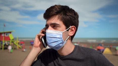 Junger-Mann-Beantwortet-Einen-Anruf-Am-Strand,-Während-Er-Aufgrund-Einer-Pandemie-Eine-Gesichtsmaske-Trägt