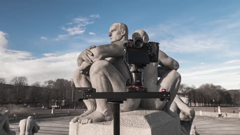 Compact-Camera-On-Slider-Shooting-Granite-Sculpture-Of-Two-Men-Sitting-Back-To-Back-With-Crossed-Arms-On-Pedestal-At-Frogner-Park,-Oslo,-Norway