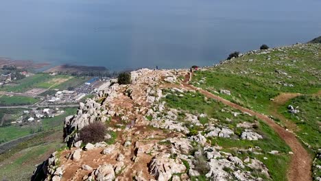Aerial-forward-shot-of-few-people-on-a-mud-trail-at-the-Arbel-cliff,-Galilee-sea-with-sun-rays-in-the-background