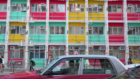 Ein-Taxi-Steht-Auf-Der-Straße,-Während-Fußgänger-An-Einem-Farbenfrohen-Sozialwohnungsgebäude-In-Hongkong-Vorbeigehen