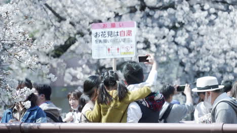 Familie-Macht-Ein-Selfie-Auf-Einem-überfüllten-Hanami-Fest-Mit-Einem-Schild,-Auf-Dem-Die-Menschen-Aufgefordert-Werden,-Inmitten-Der-Pandemie-In-Tokio,-Japan,-Zu-Hause-Zu-Bleiben