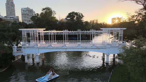 Static-shot-of-tourists-paddling-paddleboat-through-bridge-followed-by-a-flock-of-waterfowls-in-tranquil-afternoon-with-big-bright-sun-setting-at-the-background-at-rose-garden-Parque-Tres-de-Febrero