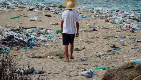 Young-man-walks-on-beach-polluted-by-plastic-junk-looking-at-industrial-facility