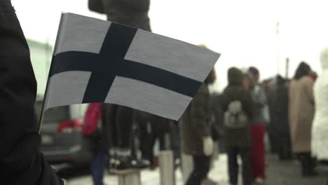 Closeup-shot-of-a-Finland-flag-being-held-by-a-protester-in-Helsinki,-crowd-of-people-in-the-background,-snowing