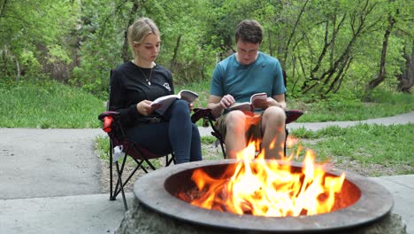 Two-Adults-Reading-Books-by-Campfire,-Outdoor-Static-Shot