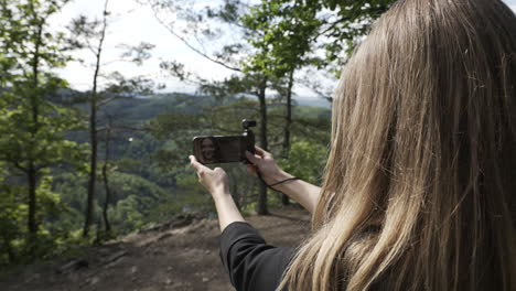 Back-of-young-woman-recording-selfie-video-with-small-gimbal-camera-and-smartphone-in-forest-on-sunny-day